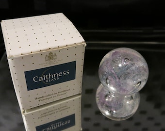 May include: A clear glass sphere with purple and gray internal marbling, reflecting on a dark surface. Beside it is a white box with gold polka dots and the text "Caithness Glass, Handcrafted in Scotland".