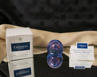 May include: A Caithness Glass miniature paperweight with a blue and red swirl design. The paperweight is displayed with its original box and a card from the Paperweight Collectors' Society. The box reads "Handcrafted in Scotland".
