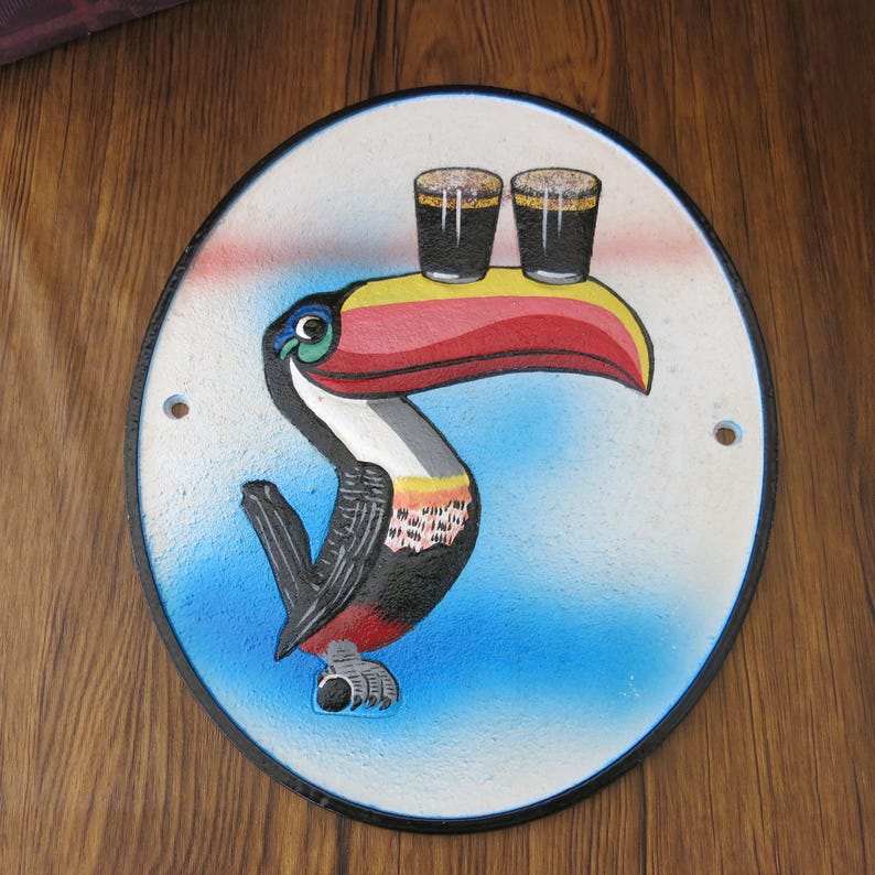 May include: A round, white, cast iron sign with a black border. A colorful toucan is painted on the sign, holding two glasses of dark beer in its beak. The background is blue.