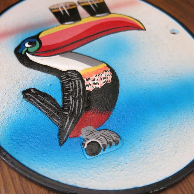 May include: A colorful toucan with a long, red beak holding three glasses of Guinness beer. The toucan is painted on a round, white and blue background.