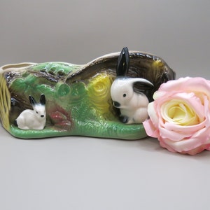 May include: A ceramic planter shaped like a tree stump with two white rabbits with black ears and eyes. The planter is painted with green, brown, and yellow colors.