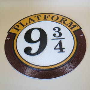 Può includere: Cartello circolare con la scritta "PLATFORM" in lettere dorate sul bordo superiore. Il numero "9 3/4" è al centro, con carattere nero su sfondo bianco, circondato da un bordo marrone.