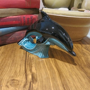 Könnte beinhalten: Eine schwarze und türkisfarbene Delfinfigur aus Keramik, die aus dem Wasser springt. Der Delfin ist glänzend und hat eine detaillierte Wellenbasis.