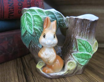 May include: A ceramic planter shaped like a tree trunk with a rabbit figurine sitting on a branch. The planter is painted brown with green leaves and has a white interior.