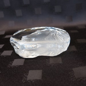 May include: A clear, oval-shaped glass sculpture with etched details. The sculpture has a frosted appearance and features a design of a face. The glass is thick and has a smooth, rounded shape. The sculpture is a decorative art piece.