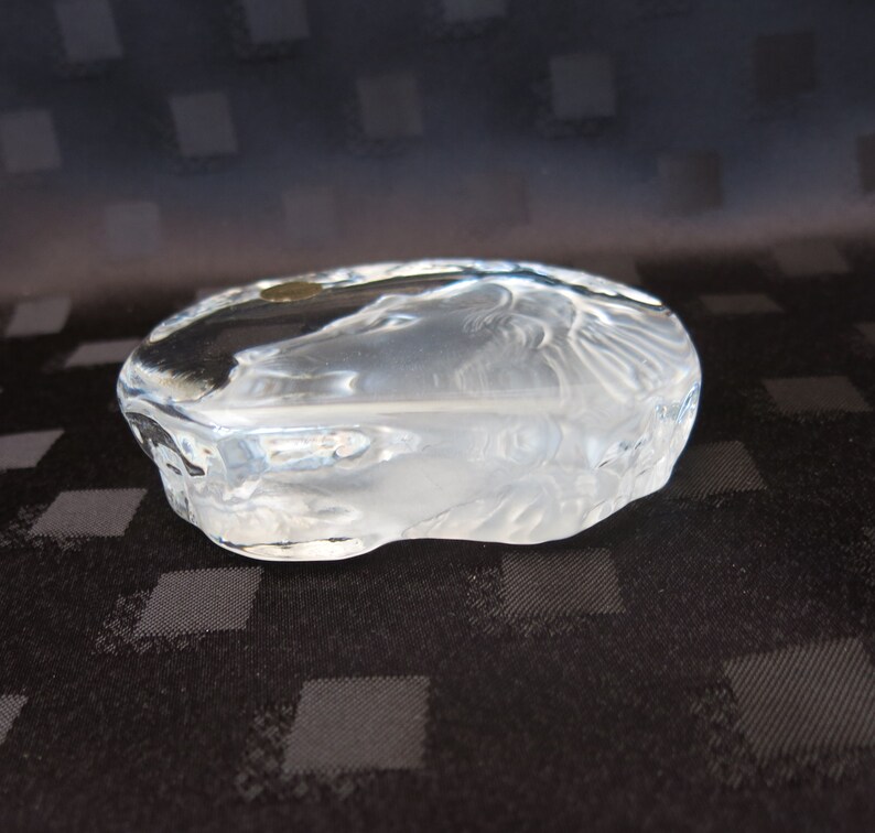 May include: A clear, oval-shaped glass sculpture with etched details. The sculpture has a frosted appearance and features a design of a face. The glass is thick and has a smooth, rounded shape. The sculpture is a decorative art piece.
