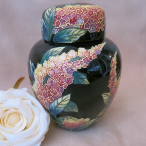 Peut inclure: Un pot en céramique noir avec un couvercle, orné d'un motif floral dans les tons de rose, jaune et vert. Le pot a une finition brillante et une forme arrondie. Une rose blanche est au premier plan.