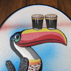 May include: A colorful painted toucan with a red, yellow, and white beak, holding two glasses of dark liquid on top of its beak. The toucan is painted on a round, white background.