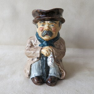Könnte beinhalten: Eine Keramikfigur eines Mannes mit braunem Hut, braunem Mantel, blauem Schal und einem Regenschirm in der Hand. Die Figur sitzt und hält ein weißes Taschentuch mit roten Punkten in der Hand.
