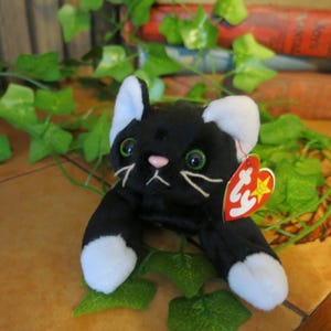 May include: A black and white plush cat toy with green eyes and a pink nose. The cat has white paws and white-tipped ears. A red heart-shaped tag is attached to the cat's ear.