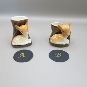 Könnte beinhalten: Zwei Keramik-Rehfiguren in Waldumgebung. Die Rehfiguren sind braun und weiß mit schwarzen Augen und sitzen auf einem braunen Sockel. Der Sockel ist wie ein Baumstumpf geformt. Die Figuren sind mit A und B gekennzeichnet.