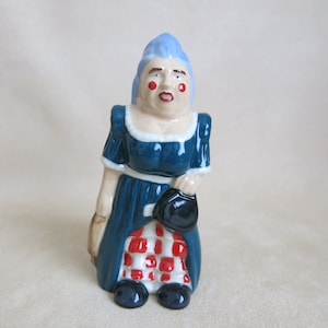 Può includere: Una statuetta in ceramica di una donna con i capelli blu, che indossa un vestito blu con una gonna a scacchi rossi e bianchi. Tiene una borsa nera nella mano destra.