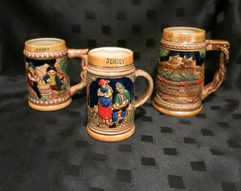 Op de afbeelding: Drie keramische bierkruiken met geschilderde scènes. De kruiken zijn bruin met blauwe en gouden accenten. De eerste kruik heeft een scène van mensen die op een veld werken. De tweede kruik heeft een scène van een stel op een veld. De derde kruik heeft een scène van een kasteel. De kruiken hebben allemaal het woord "Jersey" erop.