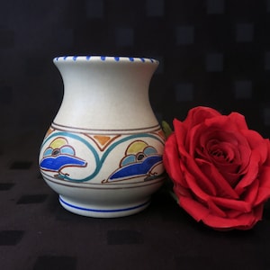 May include: A white ceramic vase with blue, orange, and yellow geometric designs. The vase is sitting on a black surface next to a red rose.
