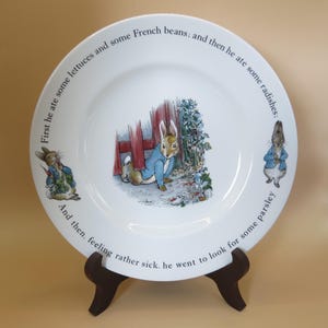 May include: White ceramic plate with illustrations of a rabbit in a blue jacket, and text around the rim. The plate depicts scenes from a story, with text that reads "First he ate some lettuces and some French beans; and then he ate some radishes:" and "And then, feeling rather sick, he went to look for some parsley."