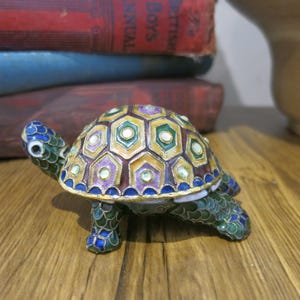 May include: A small, intricately detailed turtle figurine made of metal and decorated with colorful enamel. The turtle's shell is a mosaic of hexagonal shapes in shades of green, yellow, purple, and brown, with white accents. The turtle's legs and head are also decorated with enamel in shades of green and blue.