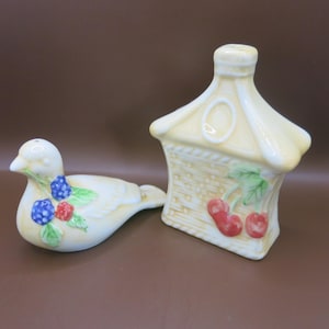 May include: A set of two ceramic salt and pepper shakers. The salt shaker is shaped like a bird with a yellow body, green leaves, and red and blue berries. The pepper shaker is shaped like a small house with a yellow roof, brown wicker walls, and red cherries.