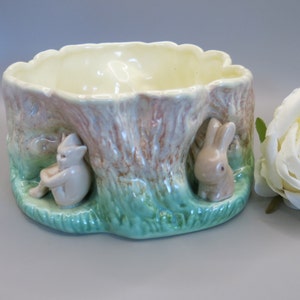 May include: A ceramic planter shaped like a tree stump with two small animal figurines, one rabbit and one cat, peeking out from the sides. The planter is painted in shades of green, brown, and pink.