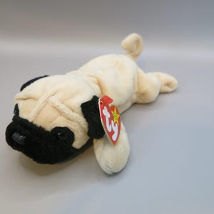 May include: A cream and black pug dog soft toy. The toy features a black snout and ears, with a cream-coloured body. A red heart-shaped tag with a yellow star is attached.