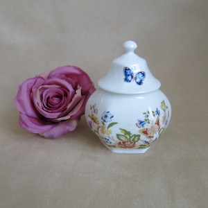 May include: A white porcelain jar with a lid, decorated with a floral pattern and a blue butterfly. The jar is sitting on a beige surface next to a pink rose.