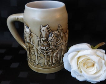 May include: A brown ceramic beer stein featuring a relief design of three horses pulling a cart. The stein has a handle and a wide mouth.