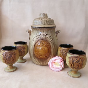 May include: A vintage ceramic rumtopf set, comprising a lidded jar and four small stemmed glasses. The jar has a raised fruit design and the word "RUMTOPE". The glasses feature a crest design. The set is in shades of brown and tan.