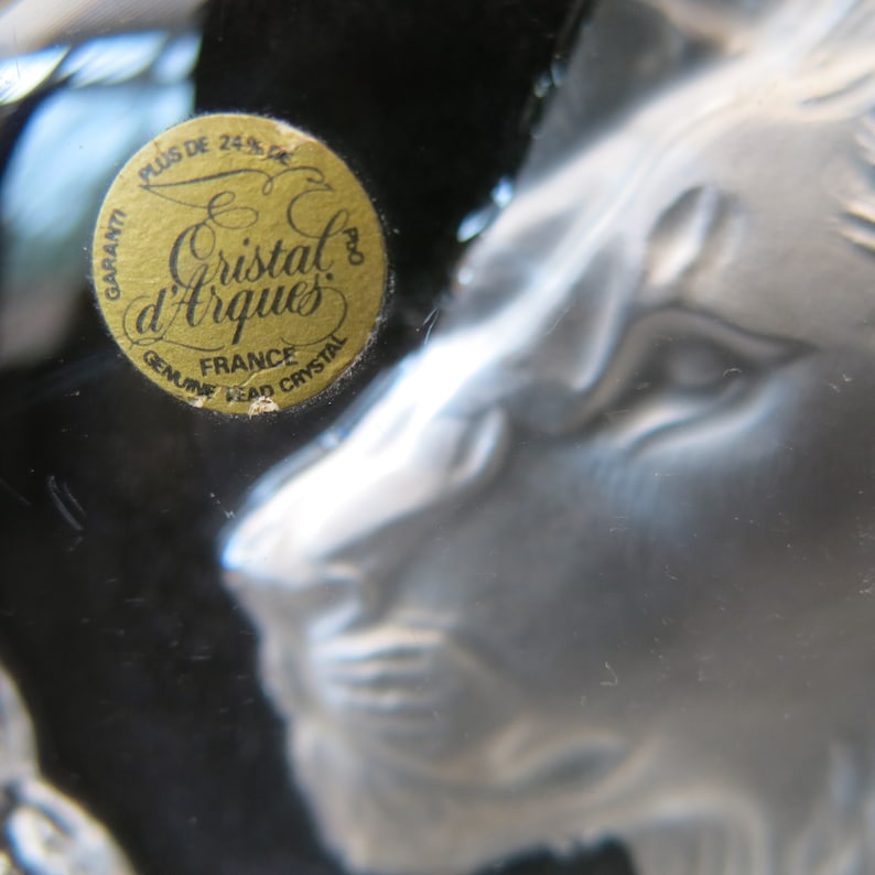 May include: Close-up of a clear crystal lion head sculpture. The sculpture has detailed features and is partially visible. A gold sticker with the text "Cristal d'Arques France Genuine Lead Crystal" is affixed to the crystal.