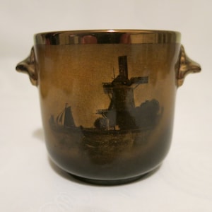 May include: A brown ceramic planter with two small handles on the sides. The planter has a gold rim and a black silhouette of a windmill and a sailboat on the front.