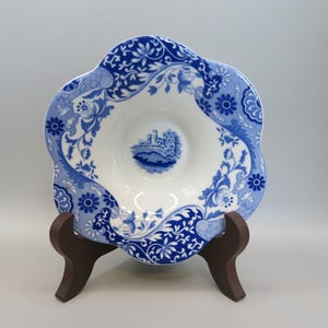 May include: A white and blue decorative bowl with a scalloped edge, featuring a floral and castle design. The bowl is supported by a brown wooden stand. The bowl's design includes floral patterns and a central castle scene.