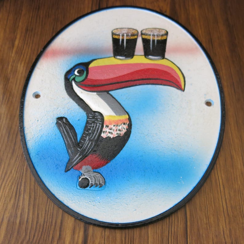 May include: A round, white cast iron sign with a black border. A toucan with a red, yellow, and black beak is holding two glasses of dark beer on its beak. The background is blue with white clouds.