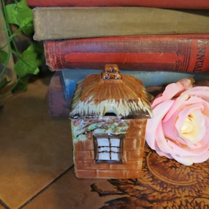 May include: A ceramic cottage-shaped container with a thatched roof and a small window. The container is brown with brick detailing and a small opening on the roof. A pink rose and antique books are in the background.