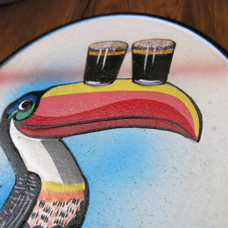 May include: A colorful toucan with a red, yellow, and black beak, holding two glasses of dark liquid on its beak. The toucan is painted on a round, white surface with a blue background.