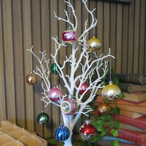 Op de afbeelding: Een witte decoratieve boom met meerdere kleurrijke kerstballen. De ornamenten zijn in verschillende kleuren, waaronder goud, rood, groen, roze en blauw. De boom staat tegen een houten achtergrond, met boeken en groen in de achtergrond.