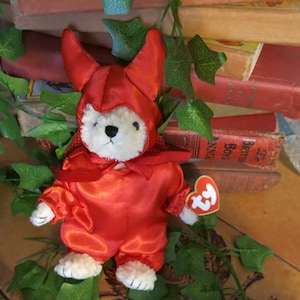 Op de afbeelding: Een pluche teddybeer gekleed in een felrood kostuum met een capuchon en cape. De beer heeft een wit gezicht en witte poten, en een hartvormig Ty-label is bevestigd. De achtergrond omvat klimop en vintage boeken.