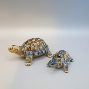 May include: Two ceramic turtle figurines. The larger turtle is in the foreground, with a brown, blue, and tan shell. The smaller turtle is behind it, with a similar color scheme. Both have detailed legs and heads, and are on a plain background.