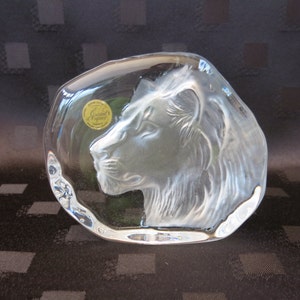 May include: A clear glass sculpture of a lion's head in profile. The lion is etched into the glass, with a detailed mane and facial features. A gold sticker with text is visible on the glass. The sculpture has a rounded, irregular shape.
