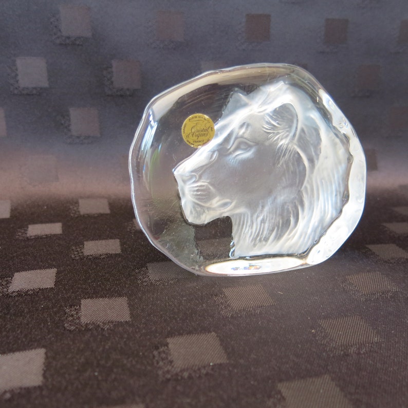 May include: A clear, oval-shaped crystal paperweight featuring a detailed etched profile of a lion's head. The paperweight has a small, round sticker with gold lettering. The lion's mane and facial features are intricately carved.