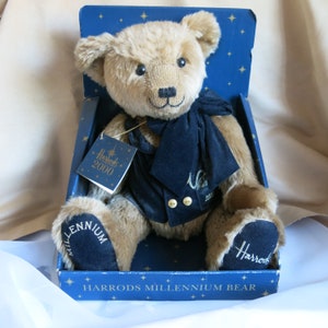 May include: A brown teddy bear wearing a navy blue velvet jacket and bow tie. The bear is sitting in a blue box with gold stars and the text "Harrods Millennium Bear" on the bottom. The bear has a tag on its chest that says "Harrods 2000".