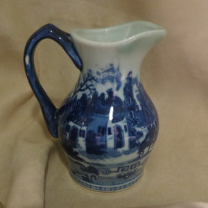 May include: A blue and white ceramic creamer with a handle and a scene of a building with trees and people in blue and white. The creamer has a blue and white border around the bottom.