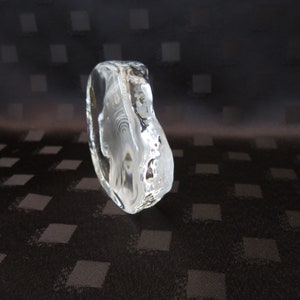 May include: A clear, sculpted glass art piece with an organic, rounded shape. The transparent object has a textured surface and reflects light, creating a shimmering effect. The background is a dark fabric with a square pattern.
