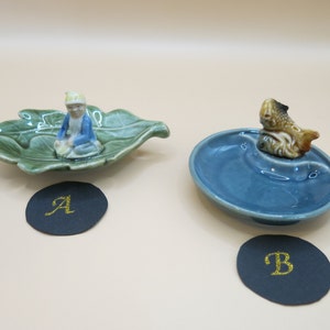 May include: Two ceramic trinket dishes. The first dish is shaped like a green leaf with a small figurine of a person sitting on it. The second dish is blue and has a small figurine of a fish on it.