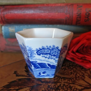May include: A white and blue ceramic hexagonal cup with a landscape design. The cup features a blue and white pastoral scene with trees, water, and animals. The cup is approximately 3 inches tall.