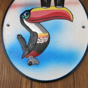 May include: A round, white and blue ceramic wall plaque with a black border. The plaque features a colorful toucan with a large, red, yellow, and black beak. The toucan is perched on a branch and is looking to the right.