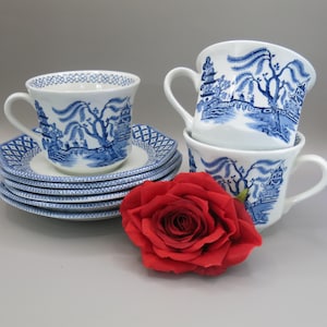 Peut inclure: Un ensemble de tasses et soucoupes à thé bleu et blanc avec un motif de saule. Les tasses et les soucoupes sont empilées les unes sur les autres, et une rose rouge est au premier plan.