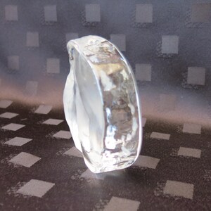 May include: A clear, sculpted glass bracelet with a textured, hammered surface. The bracelet has a rounded shape and is partially frosted, creating a play of light and shadow. The background is a dark fabric with a diamond pattern.