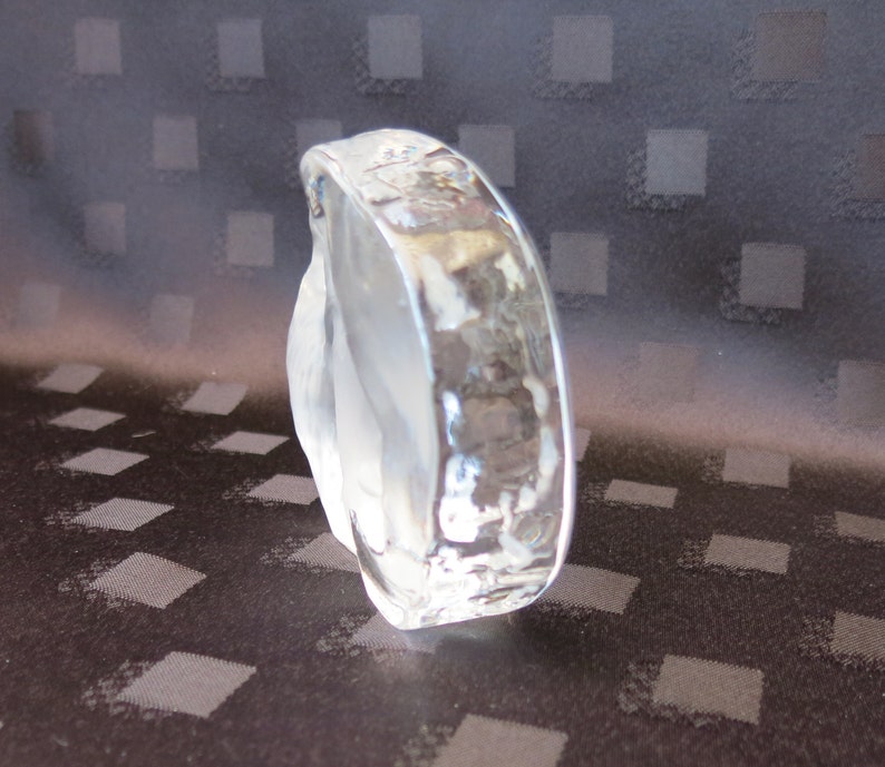 May include: A clear, sculpted glass bracelet with a textured, hammered surface. The bracelet has a rounded shape and is partially frosted, creating a play of light and shadow. The background is a dark fabric with a diamond pattern.