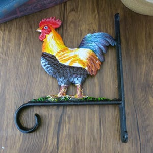 May include: A black metal rooster wall mount featuring a colourful rooster design. The rooster is painted in red, yellow, orange, blue and black. The mount has a hook on the bottom for hanging.