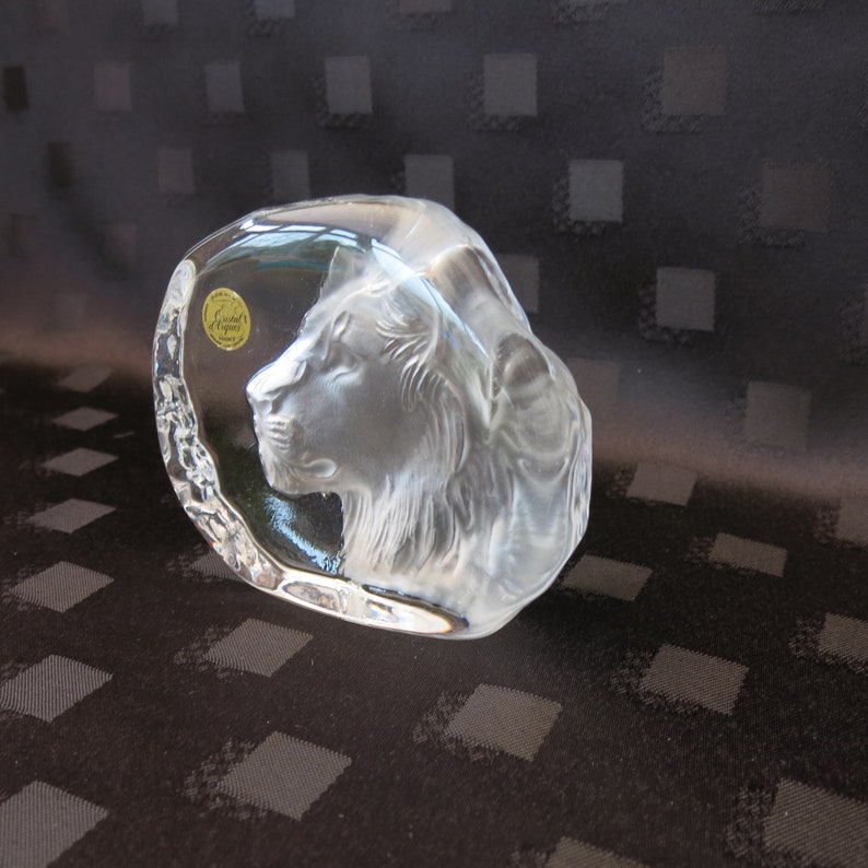 May include: A clear glass sculpture of a lion's head. The lion's profile is etched into the glass, showcasing the mane and facial features. A gold sticker with text is visible on the side. The sculpture is on a dark patterned surface.