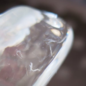 May include: Close-up of a clear glass object with a rounded edge. The glass has a slightly textured surface with visible imperfections and a subtle, reflective quality. The background is blurred, with shades of brown and gray.