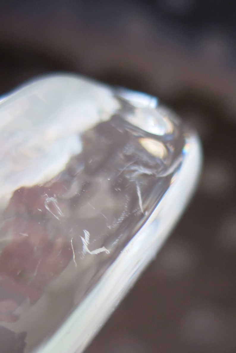 May include: Close-up of a clear glass object with a rounded edge. The glass has a slightly textured surface with visible imperfections and a subtle, reflective quality. The background is blurred, with shades of brown and gray.
